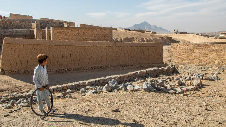 An Afghan boy plays with a stick and an inner-tube hoop outside his family's temporary home in Kabul with mountains in the background.