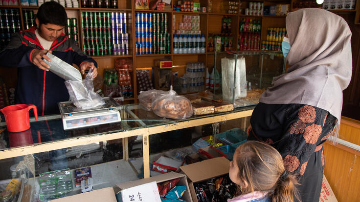 An Afghan mother and her young daughter stand at a shop counter as the shopkeeper pours bulk food into a container on a scale.