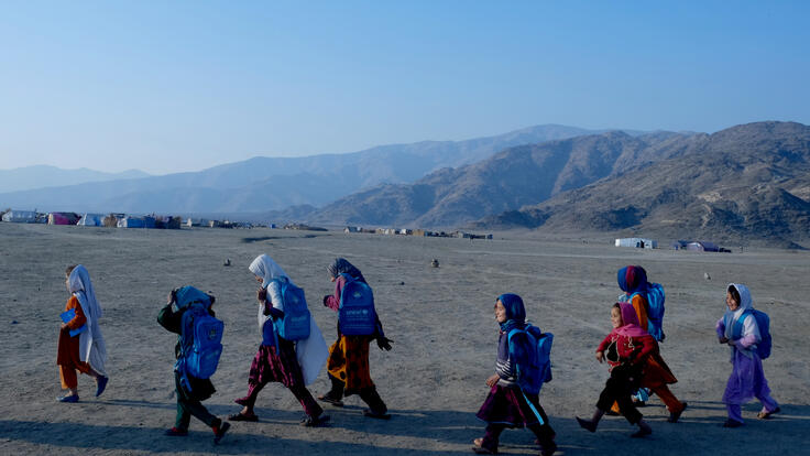 With mountains in the background, a group of girls walk past the camera all carrying backpacks. 