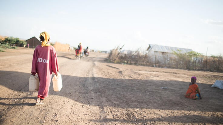 A 10-year-old girl carries two water jugs as she walks through a parched landscape near a refugee camp in Kenya.