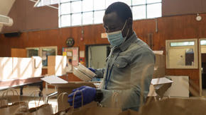 Adam Abakar, a refugee originally from Sudan, works in a food distribution center run by the IRC and World Central Kitchen. He is wearing a mask and gloves, and is putting food into paper bags. 
