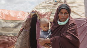 Amina holds her son while standing in front of several tents. 