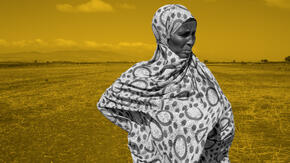 An elderly woman in Ethiopia stands in a dry field, hands at her hips.