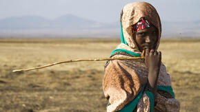A teenage girl, holding a stick and looking at the camera, stands in a drought affected very dry landscape with mountains in the distance. 