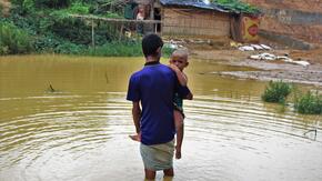 A Rohingya refugee man from Myanmar carries his two-year-old son through nearly knee-deep flood waters in a refugee camp in Bangladesh.