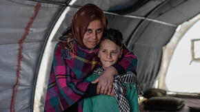 Sundus hugs her son Omar while standing behind him in a tent. Both are smiling and looking at the camera. 