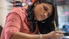 Lincy Sopall, fashion designer, sits at a desk sketching with a pencil. She was persecuted as a transgender womanin Honduras. 