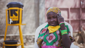 Ten-year old Anastasie sits elbow leaning on chair near an IRC hand-washing station, her COVID-19  face mask pulled down around her neck.