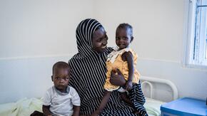 A mother sits on cot in an IRC-supported health center in Nigeria, holding her two toddler children who received care there.