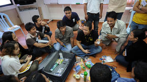 Refugees sit on the floor around an International Rescue Committee volunteer and suitcase as they learn how to strategically pack luggage.