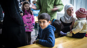 A young boy sits at a table in a refugee shelter in Serbia, surrounded by other children and their families seeking safety in Europe.