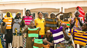 A group of women who are part of an IRC livelihoods program in Nigeria stand outside, lifting hand-knitted tops to show them off.  