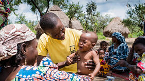 An IRC health worker holds a crying baby, giving him a medical checkup in a village in Central African Republic as his mother looks on smiling. 