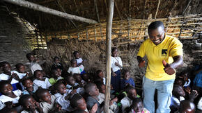 A smiling aid worker stands amid a classroom of children, leading them in a song