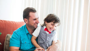 Father sits in chair inside while holding young son on his lap, both happily smiling.