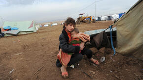 A displaced girl sits just outside a tent in Dohuk, Iraq holding a young child in her arms, bundled in a blanket.