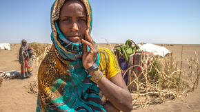 Deqa Omer, 32, stands among makeshift tents in a parched landscape in Ethiopia during a drought, her fingers resting on her cheek as she looks at the camera.