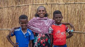A Tigrayan boy named Hafton stands smiling with his mother Wahid and his best friend Mherka outside a grass-walled shelter in a refugee camp in eastern Sudan.
