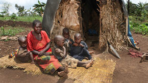 A Burundian woman and four children sit on straw mats on the the ground outside of their thatched home.