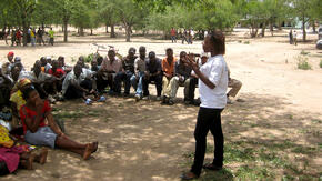 A woman who is an IRC staff member stands speaking animatedly to a group of people seated on the ground in the shade of a tree in Zimbabwe