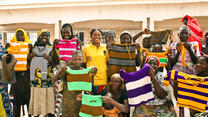 Women in Nigeria show off colorful, striped tops they've knitted to earn income as part of an IRC livelihoods program. A group of women who are part of an IRC livelihoods program in Nigeria stand outside, lifting hand-knitted tops to show them off.