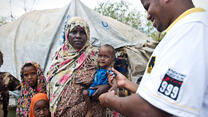 A male IRC health worker in Dadaab refugee camp stands outside a tent examining a baby girl being held in her mother's arms for signs of malnutrition.