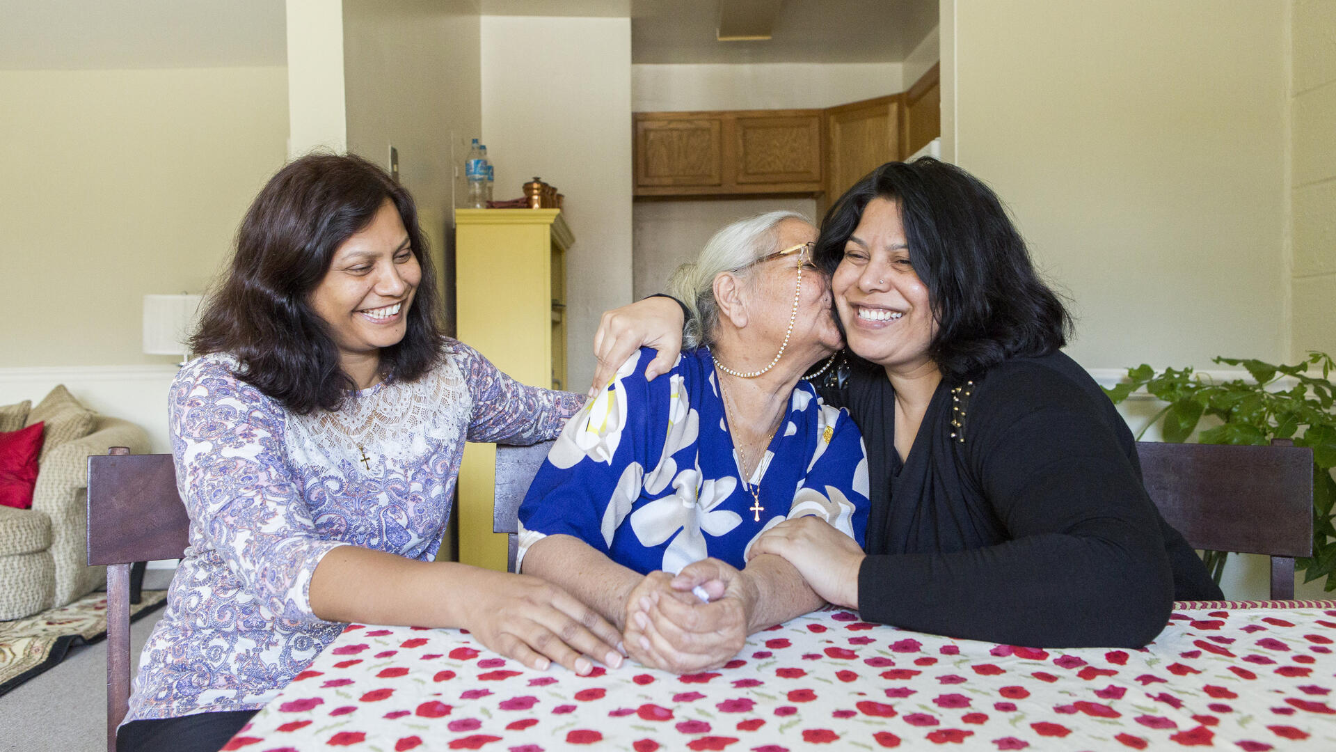 Three women in their home after coming to the U.S. through the refugee resettlement program Three women, one older and two middle aged, sit at a table with a red and white table clothe. They are smiling and the older woman is kissing the woman on her left. They are all refugees.