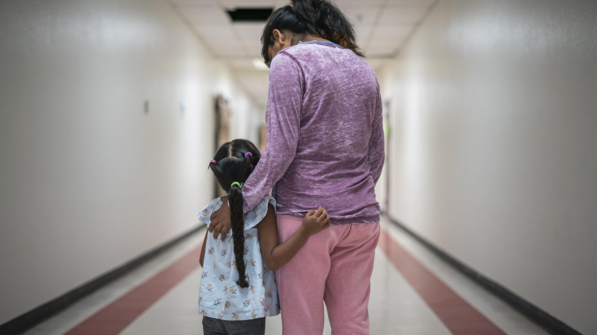 A Mexican mother wearing pink pants and a purple shirt stands with her arms around her young daughter, who has a long braid, in a hallway. They are asylum seekers in an IRC welcome center. 
