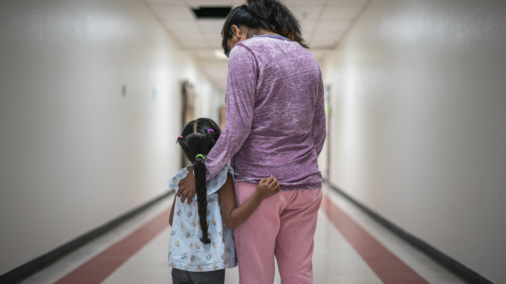 Alejandra left Mexico to seek asylum in the U.S., where she hopes to find safety for herself and her 5-year-old daughter, Ana. A Mexican mother wearing pink pants and a purple shirt stands with her arms around her young daughter, who has a long braid, in a hallway. They are asylum seekers in an IRC welcome center.