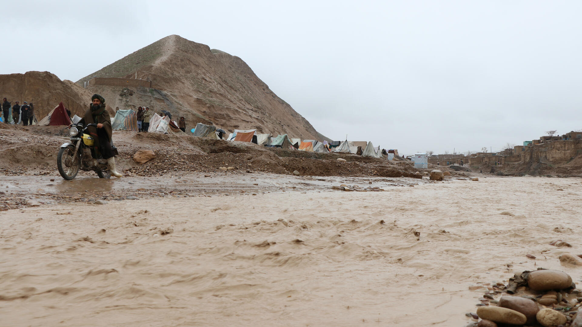 A man rides a motorcycle past muddly floodwaters in a tent camp in Afghanistan under gray skies