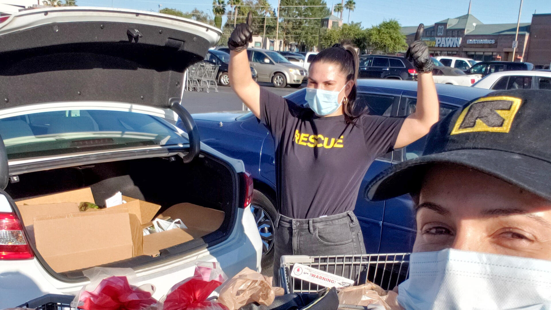 Two IRC staff members wearing IRC shirts and hats stand next to a car with an open trunk. In the trunk are groceries they recently bought for refugee and immigrant families. 