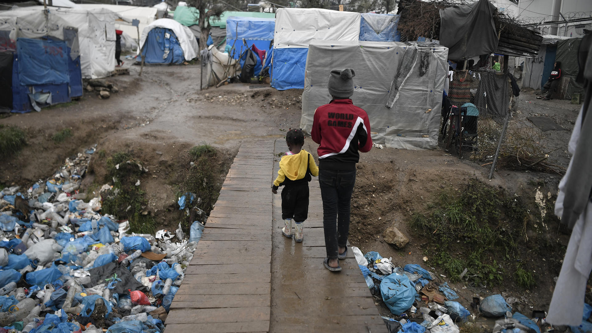 A child and a toddler walk across a wooden bridge in Moria refugee reception center, Greece.