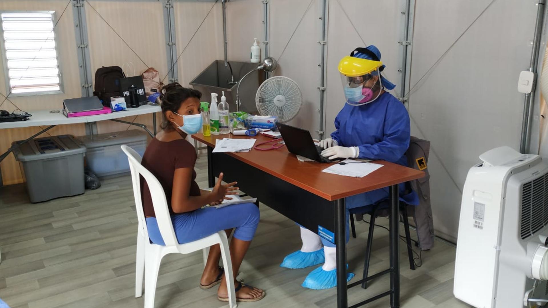 A woman receives medical care from the IRC at the Simón Bolívar bridge. A woman sits at a chair at a table with doctor in full PPE opposite her. They are in a temporary clinic set up by the IRC at the Simón Bolívar bridge.