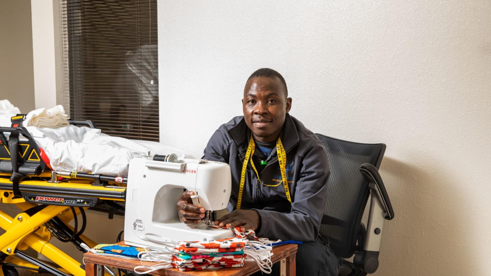 Jonathan Amissa poses with the sewing machine he uses to make masks that he is donating to the community. Jonathan Amissa sits at a desk with a sewing machine he uses to make masks for his staff and community. He is wearing yellow tape measure around his neck and is looking at the camera and smiling. There is a stack of red masks on the table.