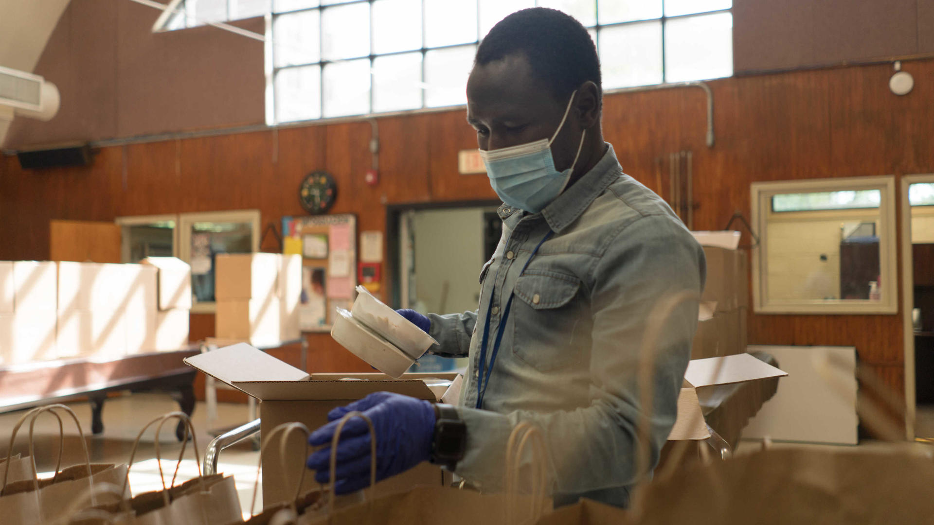 Adam Abakar, a refugee originally from Sudan, works in a food distribution center run by the IRC and World Central Kitchen Adam Abakar, a refugee originally from Sudan, works in a food distribution center run by the IRC and World Central Kitchen. He is wearing a mask and gloves, and is putting food into paper bags.