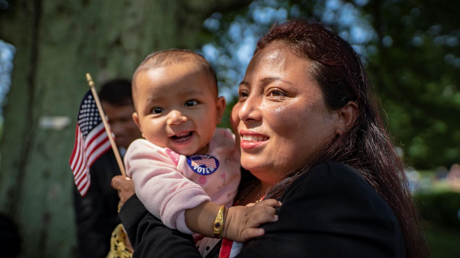 A newly naturalized citizen at a citizenship ceremony holds her baby and a small American flag. Both are smiling. 