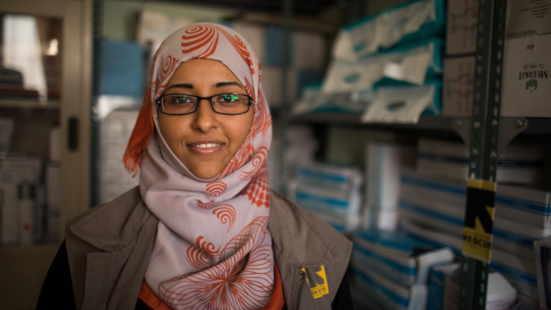 Dr. Rasha Rashed, a doctor and reproductive health manager for the IRC in Yemen. Dr. Rasha Rashed, a doctor and reproductive health manager for the IRC in Yemen, stands in a room with media supplies. She is wearing a red and white scarf and an IRC-branded vest.