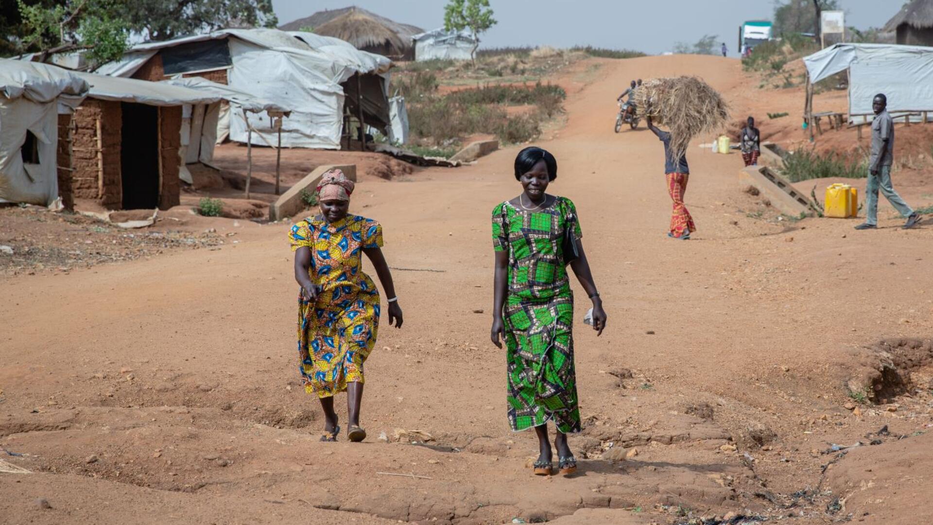 Women's rights activists Foni Grace and Loyce Tabu walk in Bidi Bidi refugeee settlement in Uganda.They are at the forefront of the photo and there are small makeshift homes and a man carrying a bale of hay behind them. 