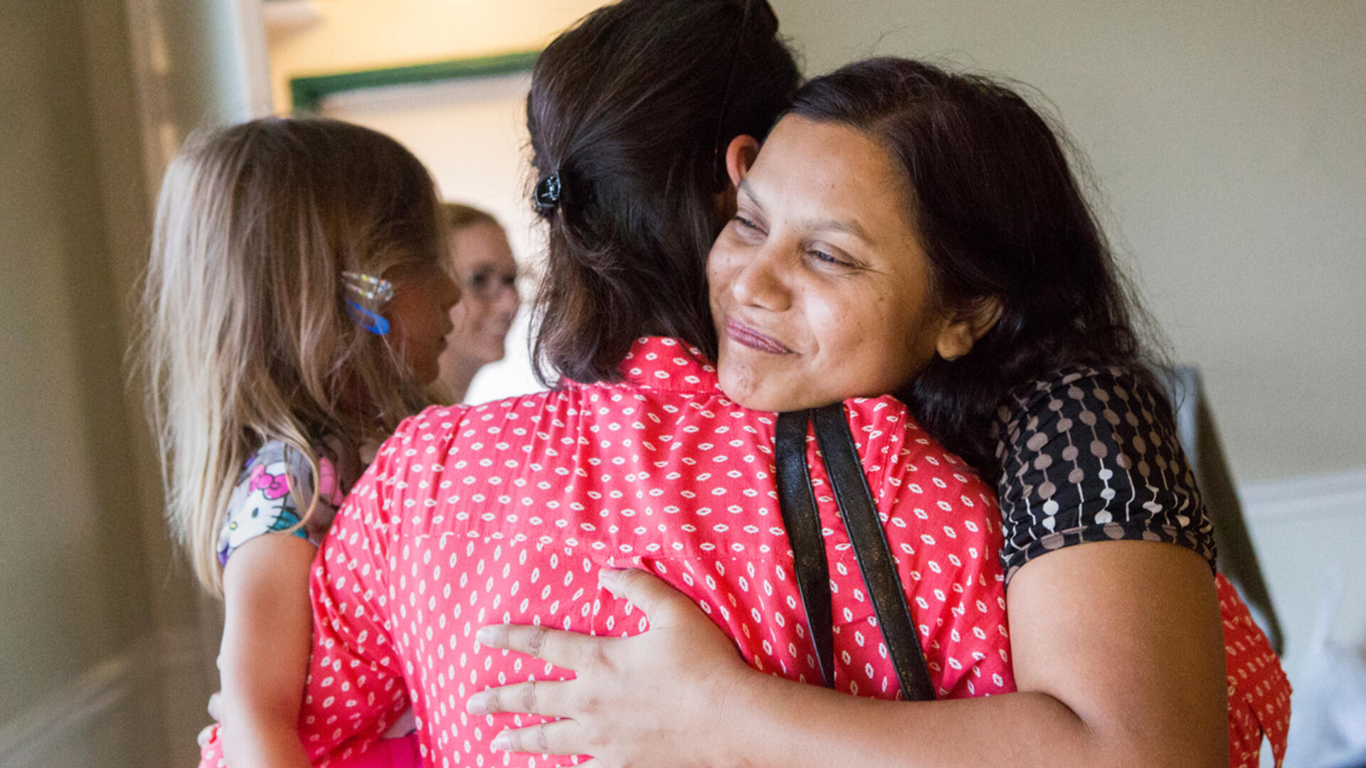 A refugee hugs her friend after safely arriving in the US Two women hug while one holds a small child. We see one of their faces, she is smiling.