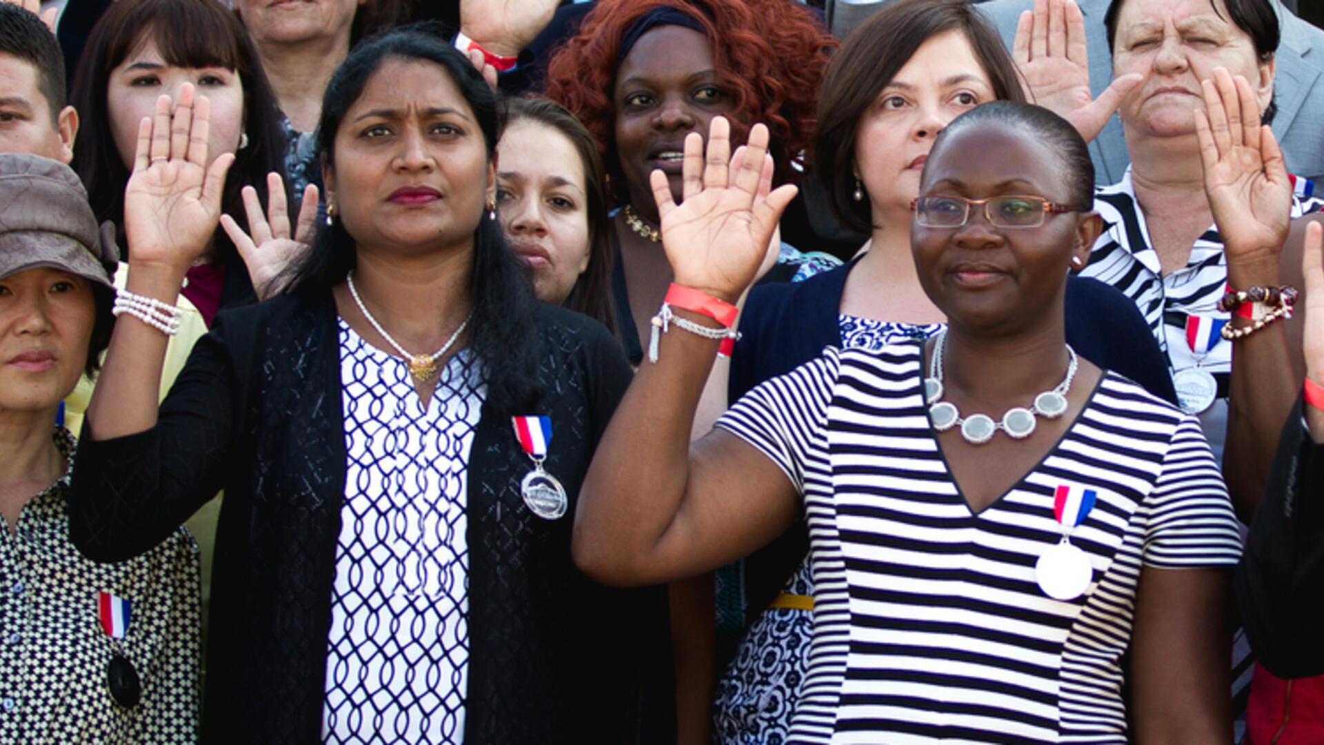 Two new citizens take their oath. They are standing in a large group of people and holding up their right hands. 