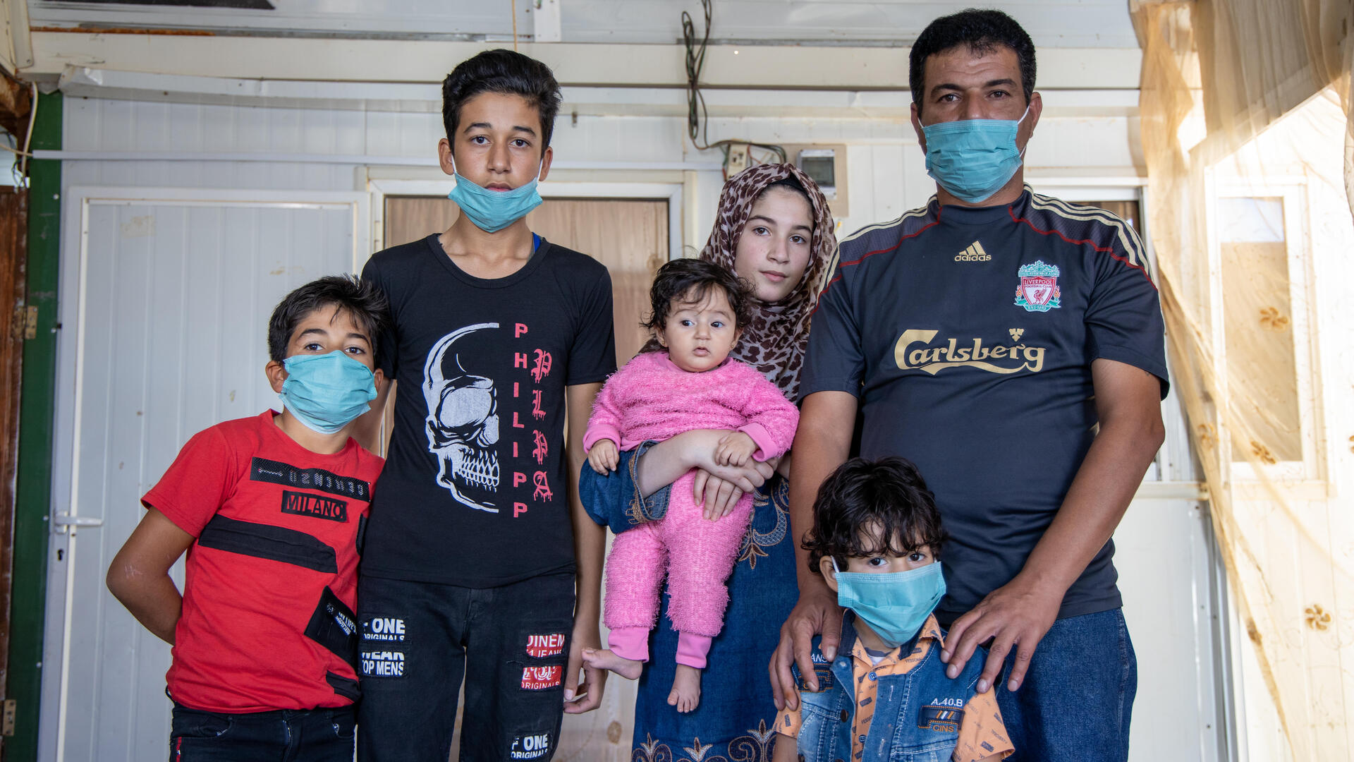 In a small tin home in a refugee camp, Manhal poses with his family: his wife holds a baby in a pink onesie, his four-year-old stands in front of him and eight-year-old and fifteen-year-old sons stands next to them. 