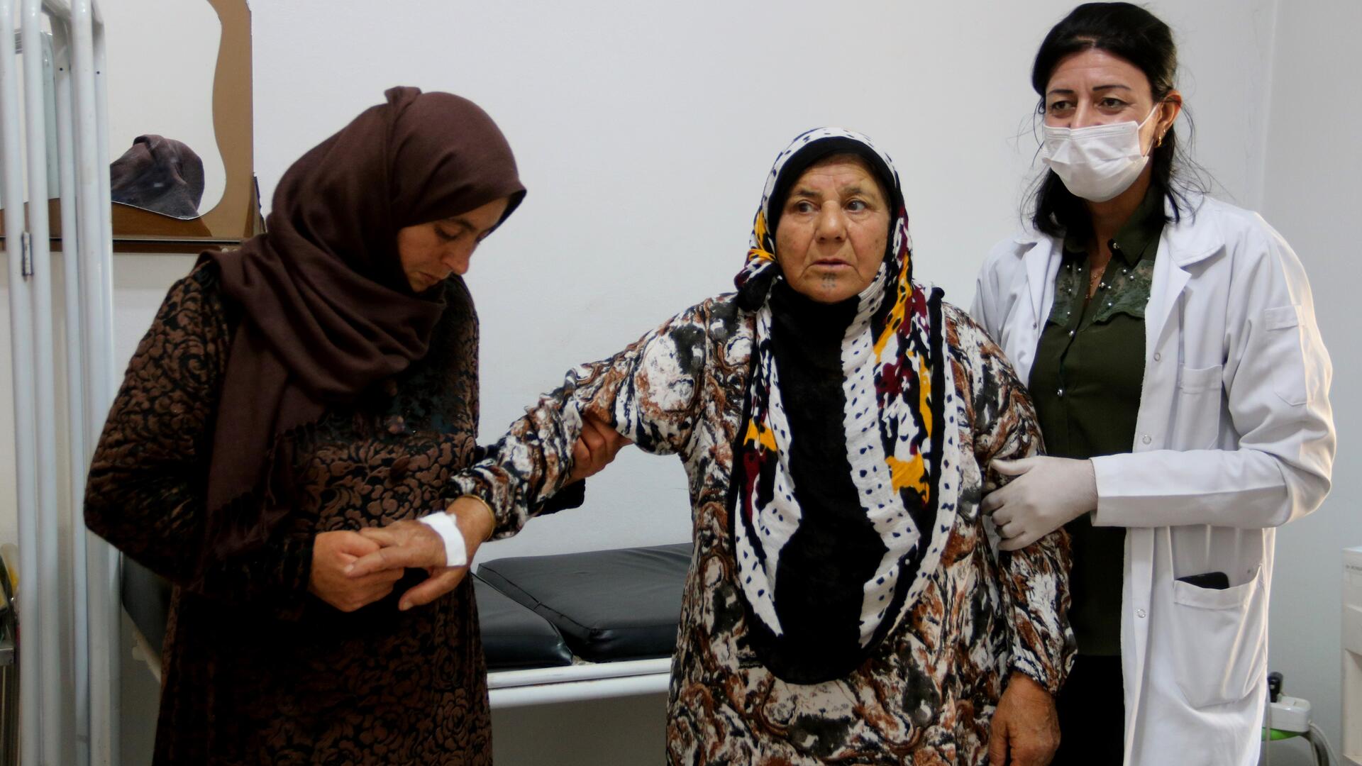 An elderly woman is supported by a relative and a doctor (names not provided) in an IRC-supported health facility in northeastern Syria. An elderly woman is assisted by a doctor and a relative in a health facility in Syria.