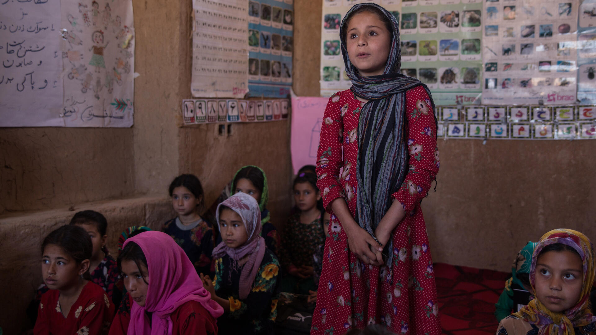 A girl in an IRC safe healing and learning space in Afghanistan A young girl stands to speak, hands clasped, in a classroom in Afghanistan.