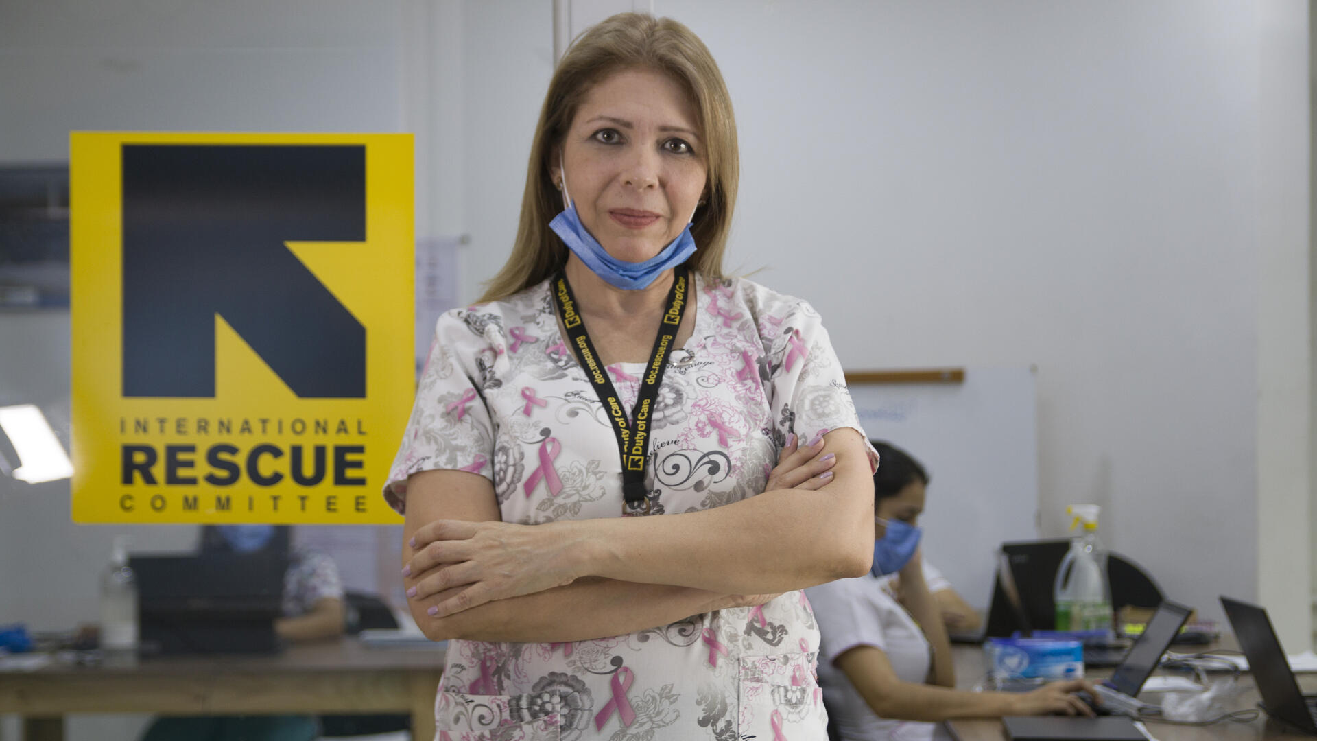 After fleeing Venezuela for Colombia, Dr. Edna Patricia Gomez worked as a doctor in an IRC clinic for sexual and reproductive health care Dr. Edna Patricia Gomez stands in scrubs with her arms crossed. She has mask at her chin and there is an IRC logo behind her.