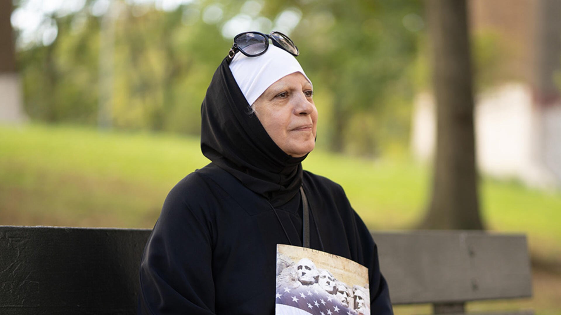 Maha al-Obaidi sits on a park bench looking off into the distance and holding a U.S. citizenship test study book. 