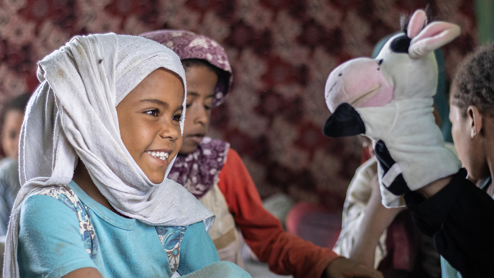 Six-year-old Ashwaq interacts with a cow puppet during an IRC-supported puppet show in Yemen A little girl smiles and looks at a cow puppet while sitting on the ground with other children in Yemen