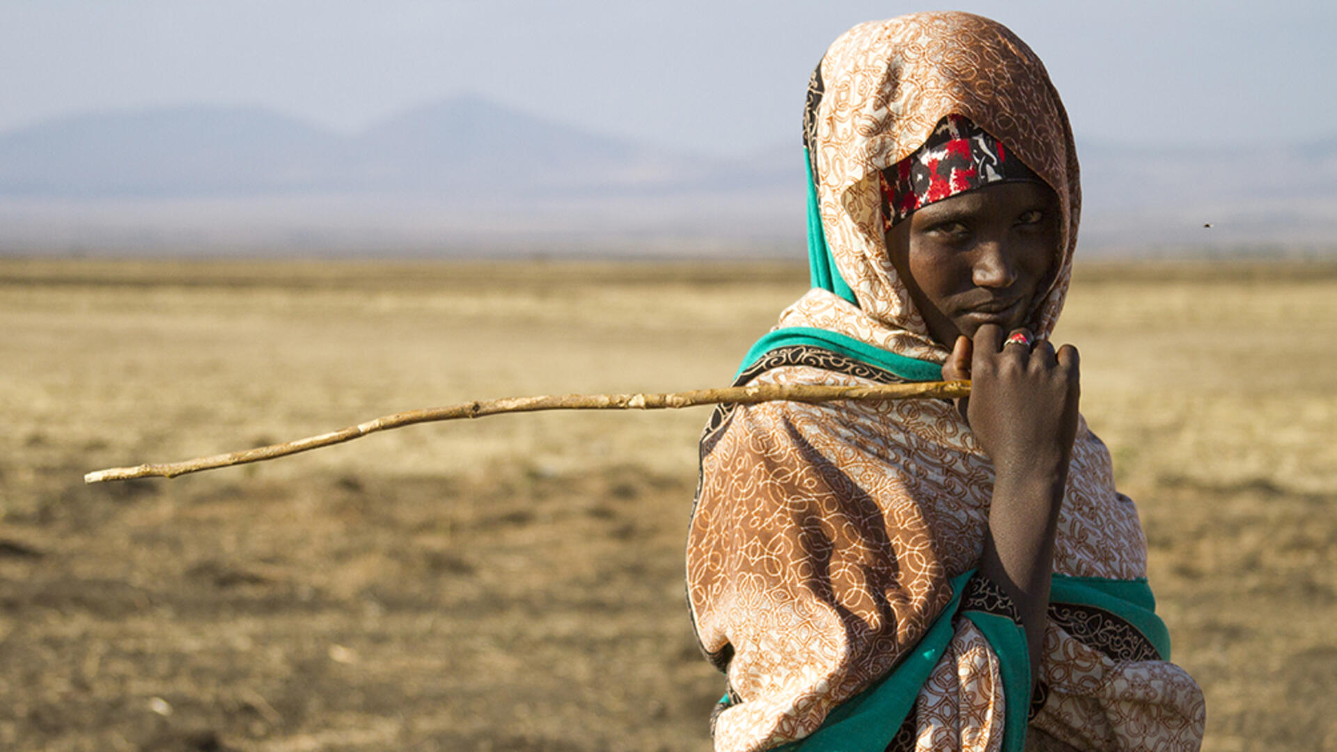 Halimo Yonis, 15, leads her donkeys to an IRC water point in the drought affected Somali region of Ethiopia. A teenage girl, holding a stick and looking at the camera, stands in a drought affected very dry landscape with mountains in the distance.