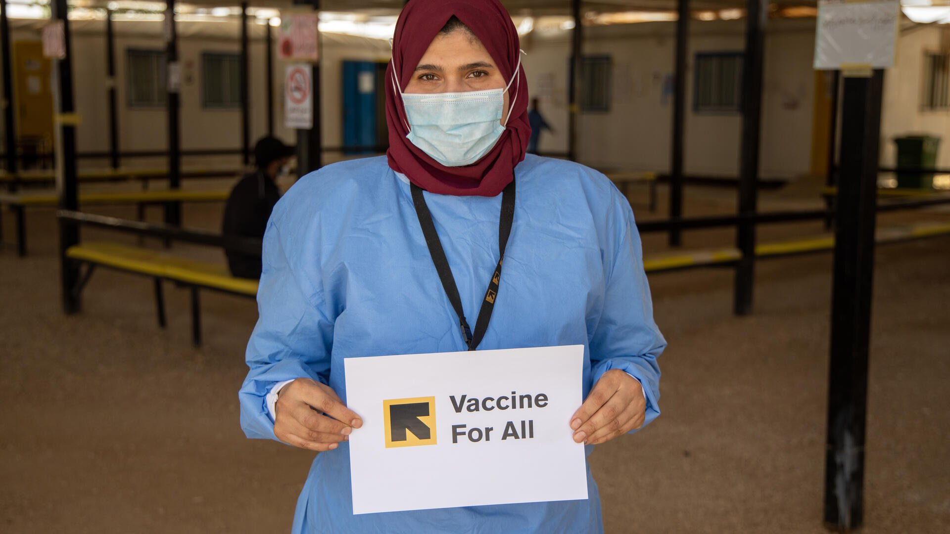A nurse wearing a mask and PPE holds a Vaccine for All sign at the IRC clinic where she works in a Syrian refugee camp in Jordan