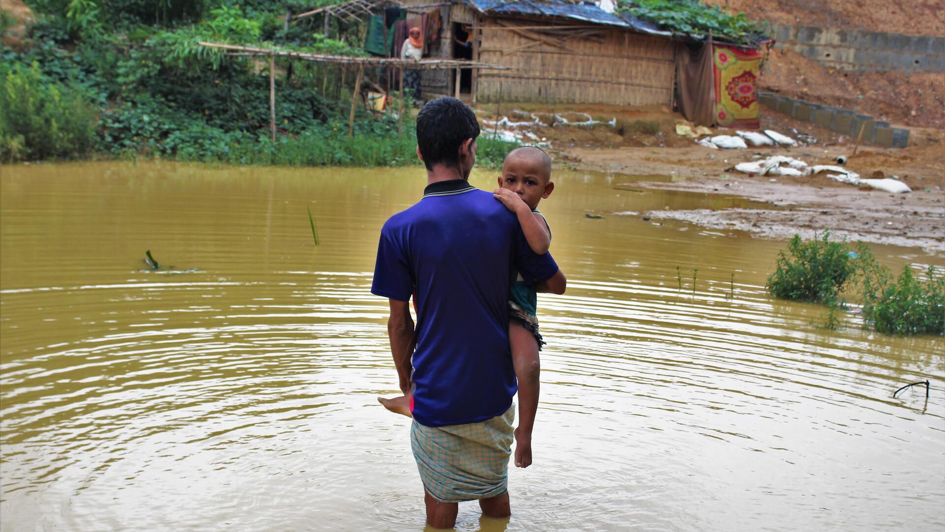 Myanmar crisis: Rohingya refugee Nurul Hauqe, 50, with his two-year-old son, wades through the monsoon-flooded Cox's Bazar refugee settlement in Bangladesh in 2019. Violence in Myanmar forced his family to flee their home in Rakhine state. A Rohingya refugee man from Myanmar carries his two-year-old son through nearly knee-deep flood waters in a refugee camp in Bangladesh.