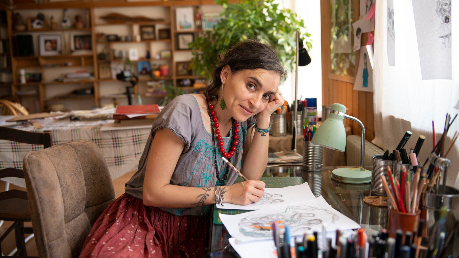 Artist and activist Diala Brisly, leaning on her desk holding a pencil, sketches an illustration of refugees for World Refugee Day 2021.